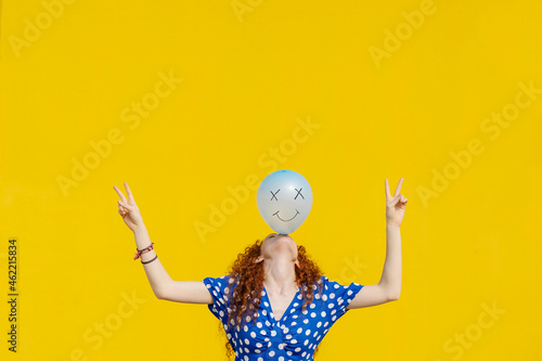 Carefree woman balancing balloon with anthropomorphic smiley face in front of yellow wall photo