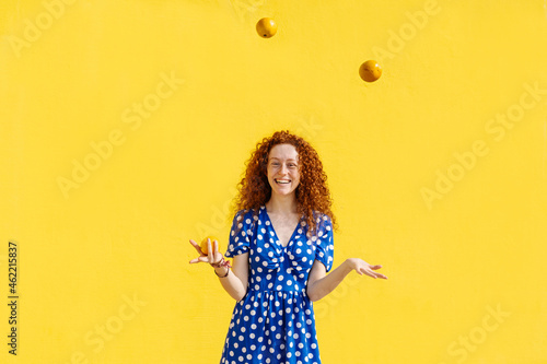 Carefree woman juggling orange fruits in front of yellow wall photo