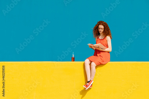 Young redhead woman reading book while sitting on yellow retaining wall