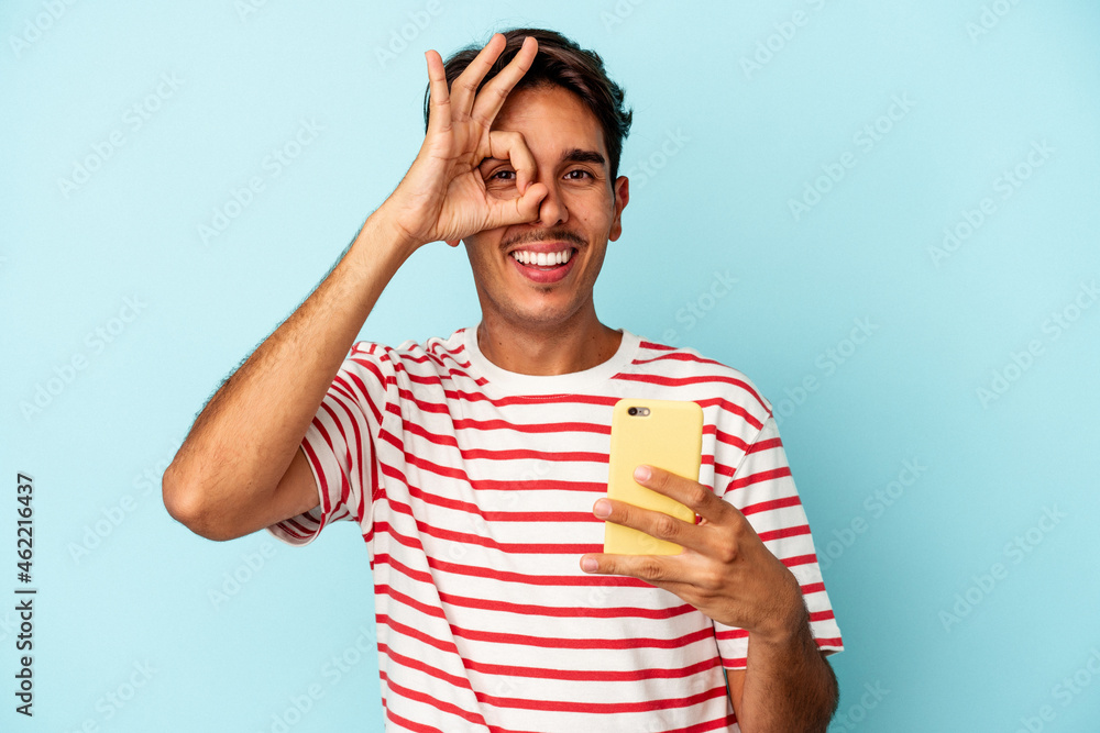 Young mixed race man holding mobile phone isolated on blue background excited keeping ok gesture on eye. Young mixed race man holding mobile phone isolated on blue background excited keeping ok gesture on eye.