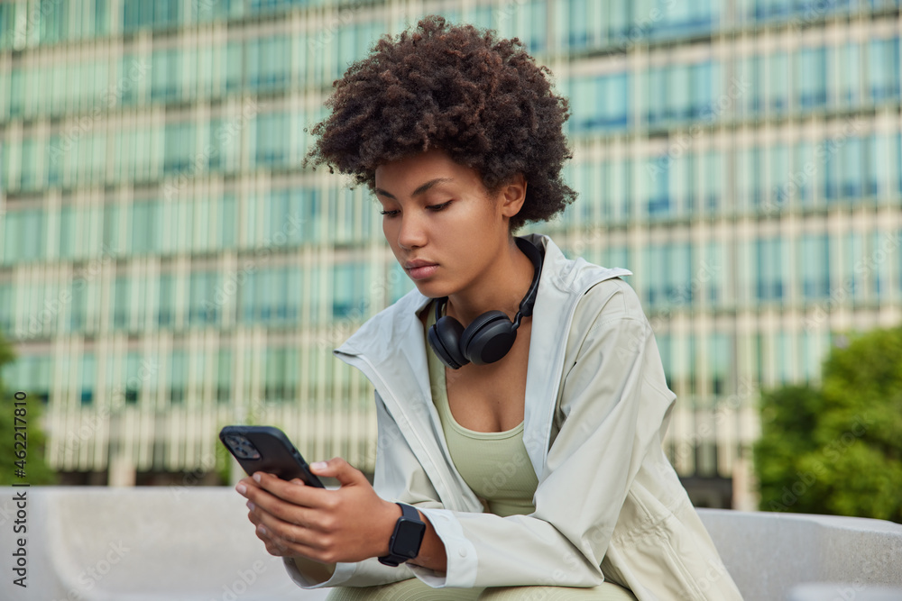 People and sporty lifestyle concept. Afro American young woman holds modern smartphone checks notification checks activity during break after workout browses internet poses against modern building - obrazy, fototapety, plakaty 