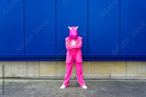 Woman wearing vibrant pink suit and hippo mask standing in front of blue wall with crossed arms photo