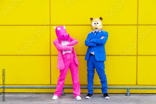 Man and woman wearing vibrant suits and animal masks posing together with crossed arms in front of yellow wall photo
