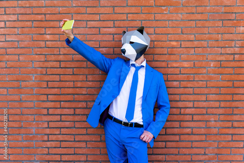 Man wearing vibrant blue suit and raccoon mask taking smart phone selfie against brick wall photo