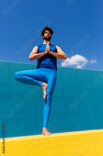 Man with hands clasped practicing Vrikshasana while standing on retaining wall photo