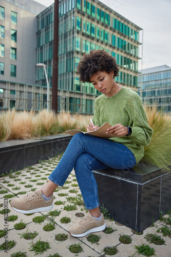 Outdoor shot of pensive Afro American woman writes down informative plan in notebook or organizer makes education notes poses in urban setting studies outside prepares for test enjoys good day