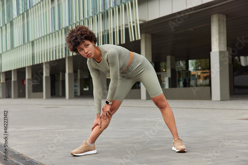Sportive young motivated African American woman leans to knees warms up before workout dressed in sportswear has determined face expression poses at downtown trains muscles has regular training
