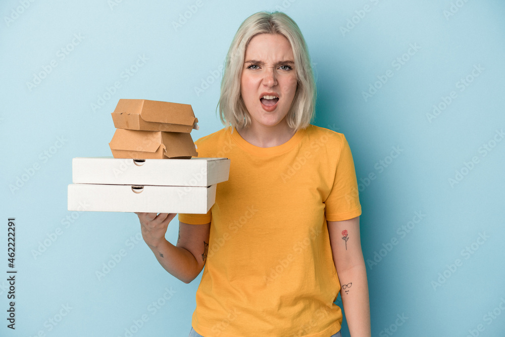 Young caucasian woman holding pizzas and burgers isolated on blue background screaming very angry and aggressive. Young caucasian woman holding pizzas and burgers isolated on blue background screaming very angry and aggressive.