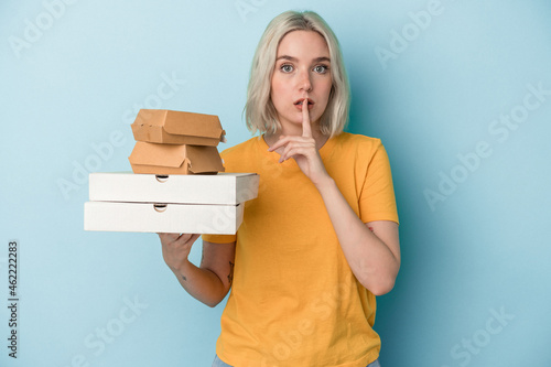 Young caucasian woman holding pizzas and burgers isolated on blue background keeping a secret or asking for silence.