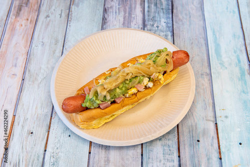 Hot dog with boiled egg, guacamole, boiled ham and onion on top on cardboard plate