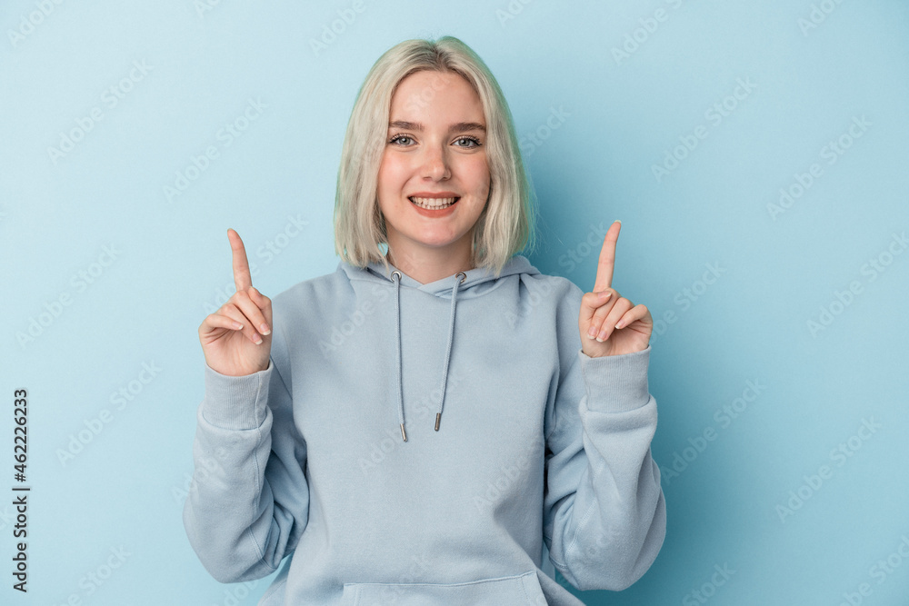 Young caucasian woman isolated on blue background indicates with both fore fingers up showing a blank space. Young caucasian woman isolated on blue background indicates with both fore fingers up showing a blank space.