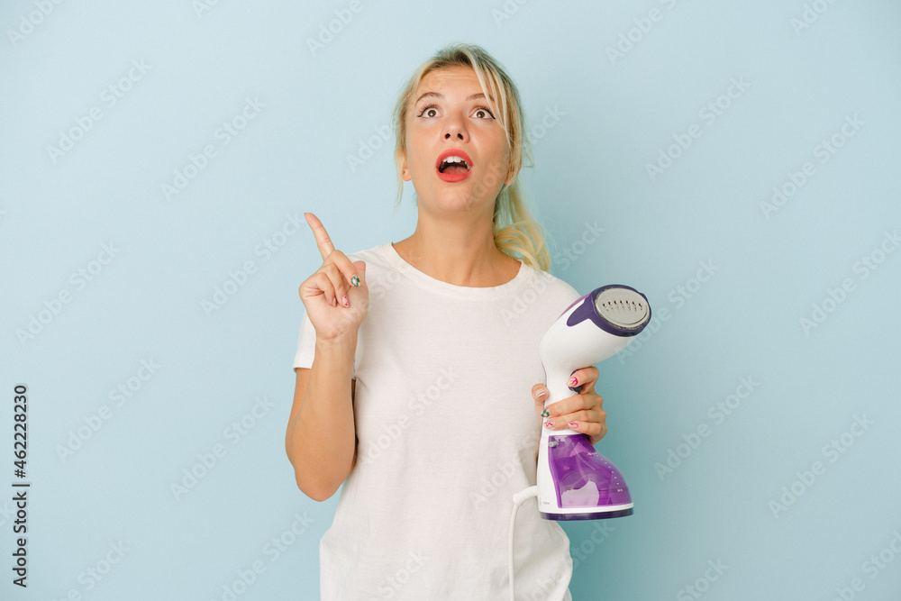 Young Russian woman holding vertical iron isolated on blue background pointing upside with opened mouth.