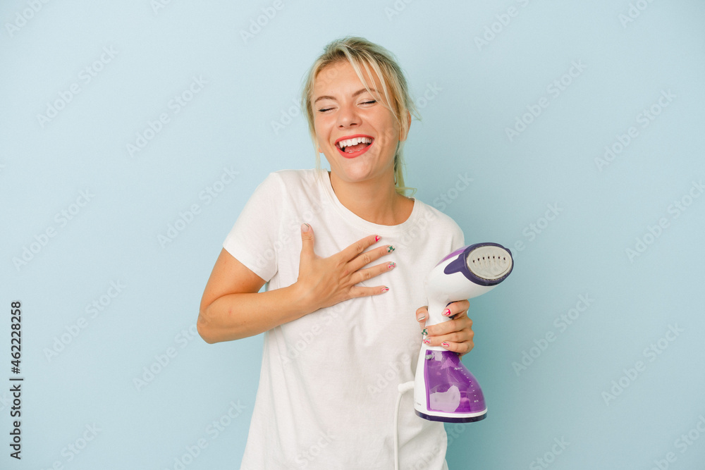 Young Russian woman holding vertical iron isolated on blue background laughs out loudly keeping hand on chest. Young Russian woman holding vertical iron isolated on blue background laughs out loudly keeping hand on chest.