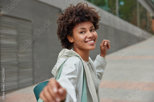 Carefree happy Afro American woman returns in good mood after fitness training carried rolled up karemat dressed in anorak looks with joyful expression poses outside against blurred background