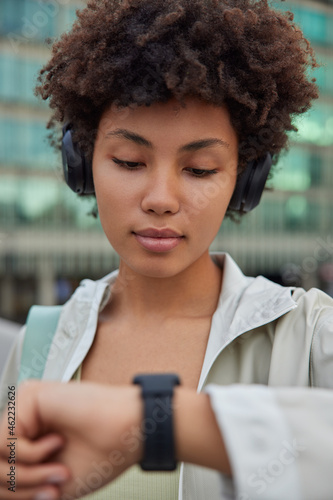 Close up shot of serious Afro American woman looks at smartwatch on wrist tracks health after training listens favorite audio track in headphones dressed in jacket poses against blurred background