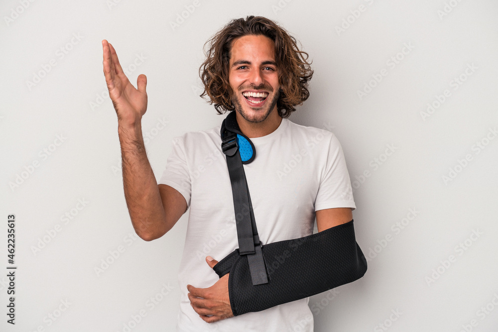 Young caucasian man with broken hand isolated on gray background receiving a pleasant surprise, excited and raising hands. Young caucasian man with broken hand isolated on gray background receiving a pleasant surprise, excited and raising hands.