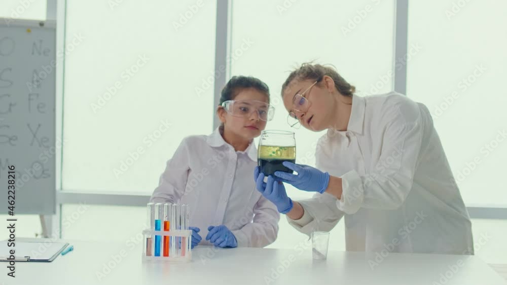 A Teenage Girl With a Teacher in Lab Coats and Safety Glasses Conducts Experiments, Studies the Color of the Reagent in Test Tubes, Takes Notes in Scientific Laboratory. Education, Early Development. A Teenage Girl With a Teacher in Lab Coats and Safety Glasses Conducts Experiments, Studies the Color of the Reagent in Test Tubes, Takes Notes in Scientific Laboratory. Education, Early Development.