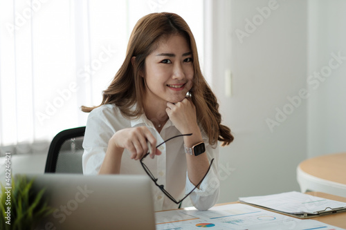 Portrait young beautiful smiling asian business woman holding a glasses and laptop Placed at the wooden table at the office