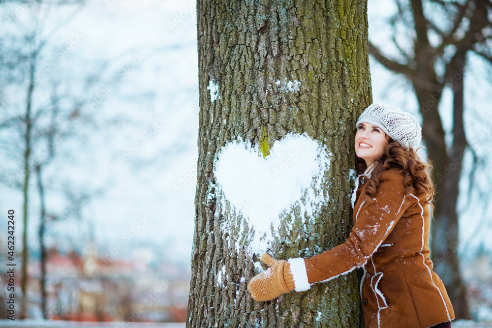 smiling stylish woman hugging tree with snowy heart - obrazy, fototapety, plakaty 