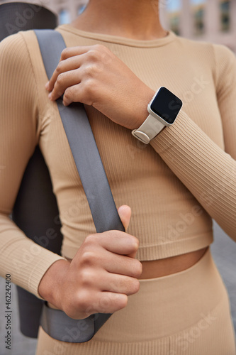 Cropped shot of unrecognizable slim woman dressed in beige tracksuit carried rolled fitness mat wears smartwatch ready for training. Faceless female instructor going to conduct yoga lessons.
