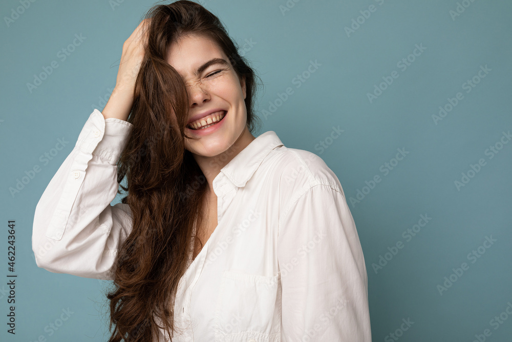 Portrait of beautiful positive cheerful cute smiling young brunette woman in casual white shirt isolated on blue background with copy space Portrait of beautiful positive cheerful cute smiling young brunette woman in casual white shirt isolated on blue background with copy space