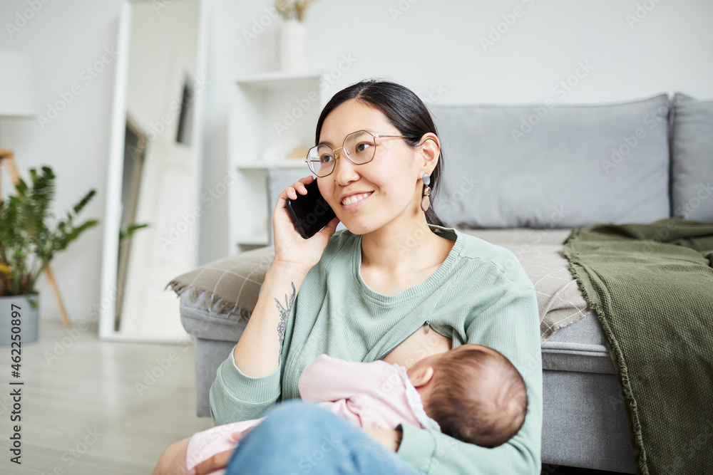 Happy Asian mother talking on mobile phone while feeding her baby sitting in the living room Happy Asian mother talking on mobile phone while feeding her baby sitting in the living room