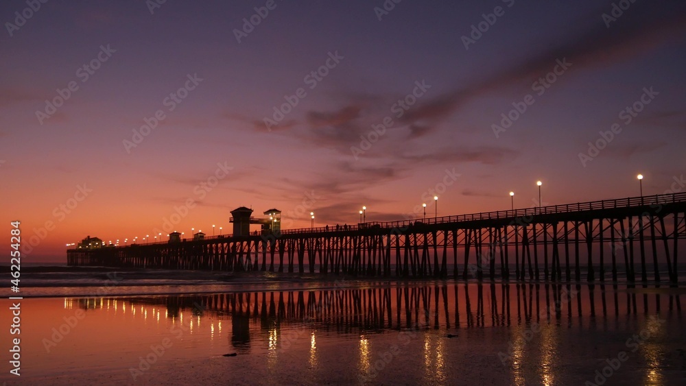 Fototapeta premium Pier silhouette Oceanside California USA. Pacific ocean tide tropical beach. Summertime gloaming atmosphere. Purple aesthetic gradient, calm twilight sky, pink violet dusk. Lights reflection in water.