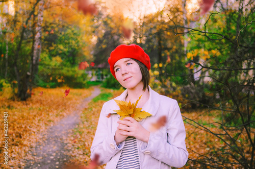 Happy girl is wearing white coat and red beret in park with yellow leaves. Fall time. Close-up portrait of pretty french girl resting in autum park