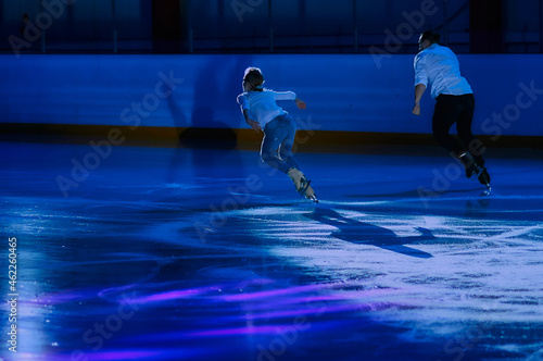A pair of skaters glide across patterned ice performing shows or competitions in the ice arena. A man and a woman are illuminated by a spotlight. Detailed ice texture. Selective focus. Motion blur.