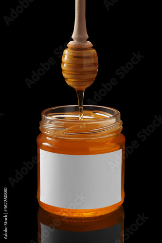 Honey dripping from dipper into full glass jar with white blank label isolated on black background. Product mock up.