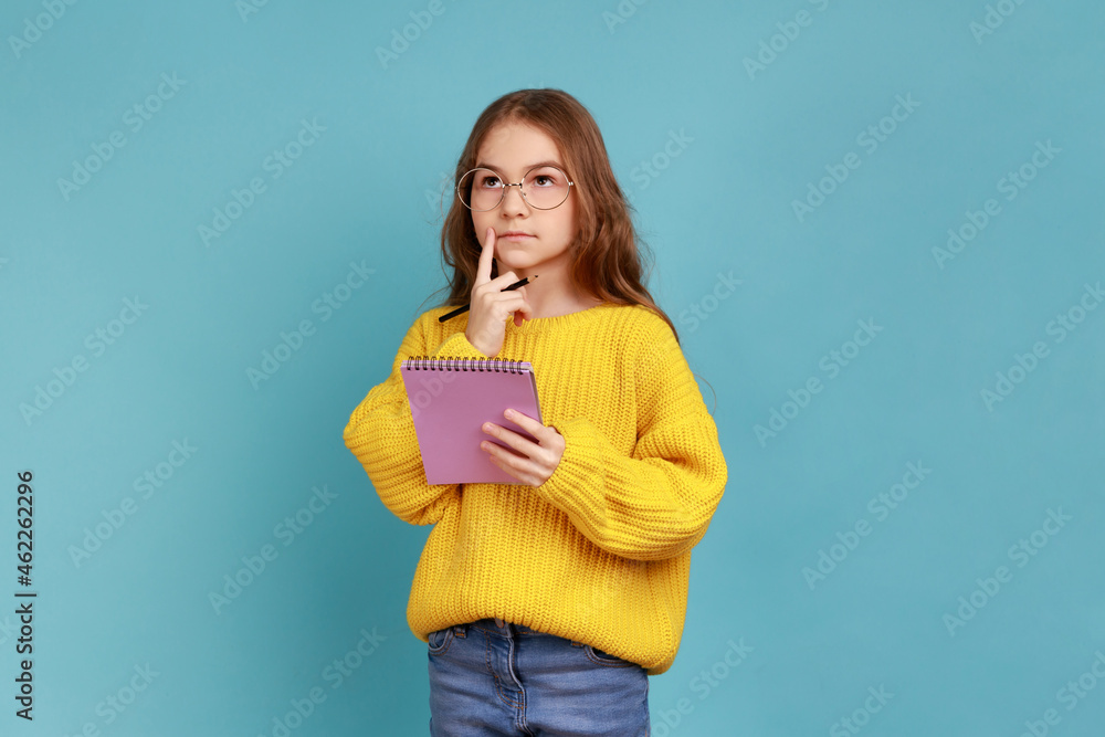 Portrait of little girl writing in notebook, thinking, taking notes, school child doing homework, wearing yellow casual style sweater. Indoor studio shot isolated on blue background. Portrait of little girl writing in notebook, thinking, taking notes, school child doing homework, wearing yellow casual style sweater. Indoor studio shot isolated on blue background.
