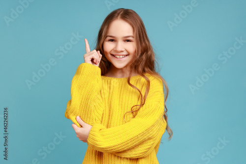 Portrait of little girl points finger up  looking inspired by genius thought  showing good idea sign  wearing yellow casual style sweater. Indoor studio shot isolated on blue background.