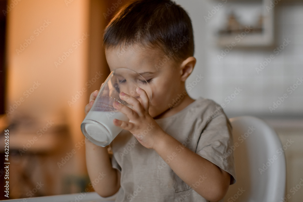 A three-year-old little boy drinks milk from a glass in the kitchen through a paper tube A three-year-old little boy drinks milk from a glass in the kitchen through a paper tube