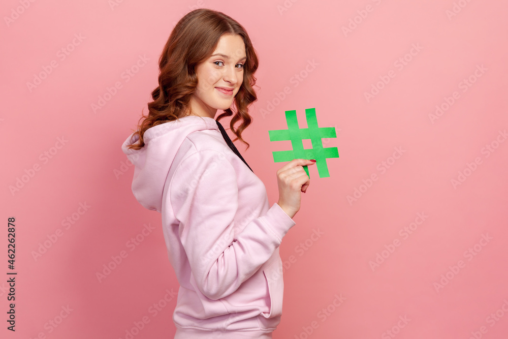 Side view portrait of happy curly-haired teen female in hoodie showing hashtag sign, meaning blog recommendation, tagging viral post. Indoor studio shot isolated on pink background Side view portrait of happy curly-haired teen female in hoodie showing hashtag sign, meaning blog recommendation, tagging viral post. Indoor studio shot isolated on pink background