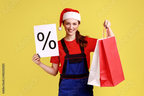 Positive courier female shows paper with percent mark, shows shopping bags, discounts for delivering, wearing blue overalls and santa claus hat. Indoor studio shot isolated on yellow background.