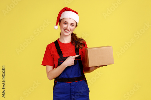 Pleased delivery woman holding pointing at parcel, fast delivering of presents for winter holiday, wearing blue overalls and santa claus hat. Indoor studio shot isolated on yellow background.