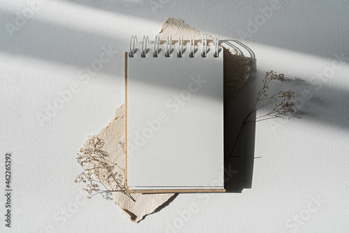 Notepad mockup for showcasing artwork and design. A notebook with an open blank page lies on a white table surrounded by dry plants. Minimal mock up of sketchbook or drawing paper. photo