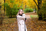 A woman with blonde hair walks in an autumn park with a cup of coffee in her hands . Golden autumn, the colors of autumn. A young woman in a light long raincoat walks through the autumn forest