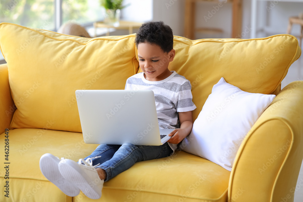 Little African-American boy with laptop watching cartoons on sofa at home
