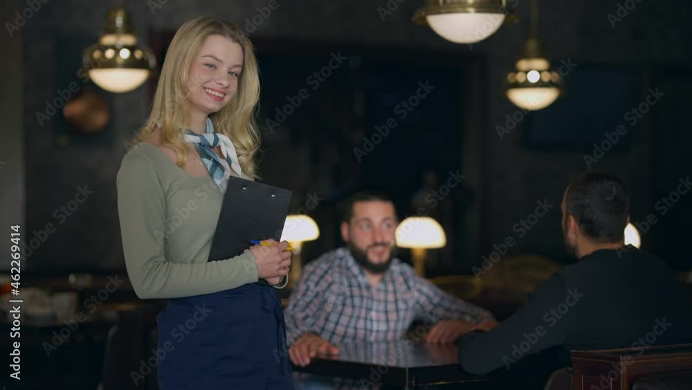 Happy smiling young waitress posing in restaurant with blurred men talking sitting table at background. Portrait of charming confident professional Caucasian waiter looking at camera standing indoors Happy smiling young waitress posing in restaurant with blurred men talking sitting table at background. Portrait of charming confident professional Caucasian waiter looking at camera standing indoors