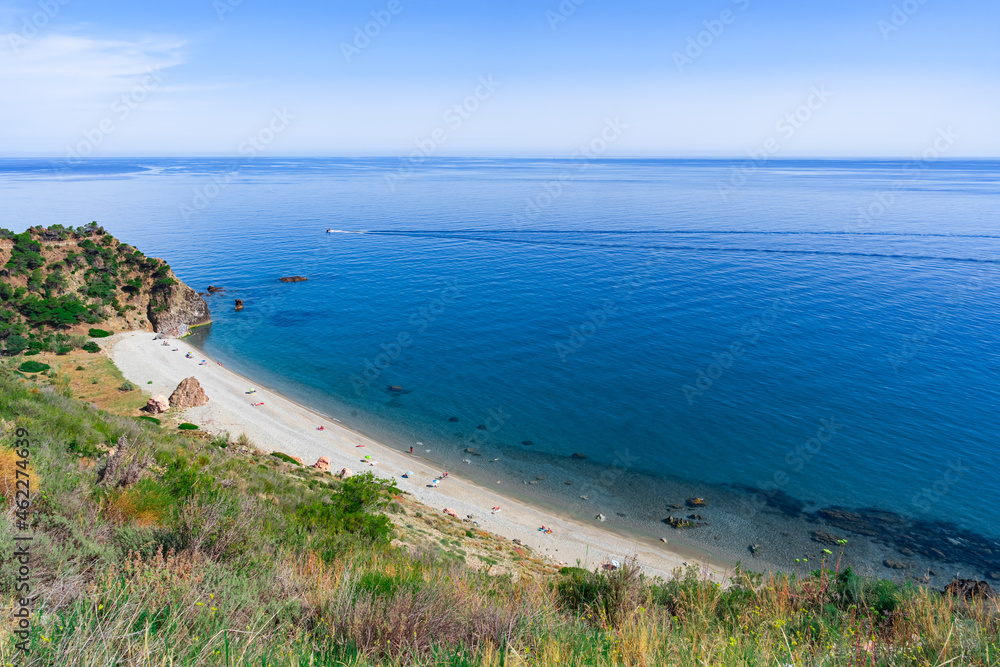 Cala salvaje con laderas y barrancos con vegetación de clima mediterráneo a su espalda y las azules y transparentes aguas del mar mediterráneo a su frente un día soleado con cielo azul.