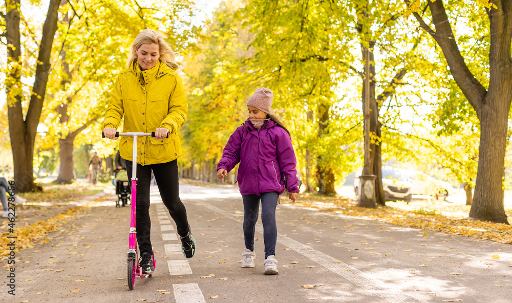 Mother and daughter are walking in the autumn in the park. woman is riding a scooter Mother and daughter are walking in the autumn in the park. woman is riding a scooter