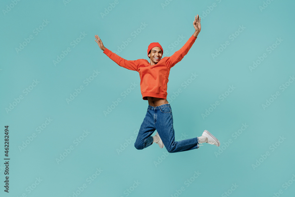 Full body fun cool overjoyed young smiling happy african american man 20s wear orange shirt hat jump high with outstretched hands laugh isolated on plain pastel light blue background studio portrait