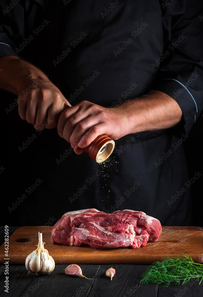 Professional chef cooking raw meat fillet and adding pepper or chili for marinade on black background. Working environment in kitchen in restaurant or cafe