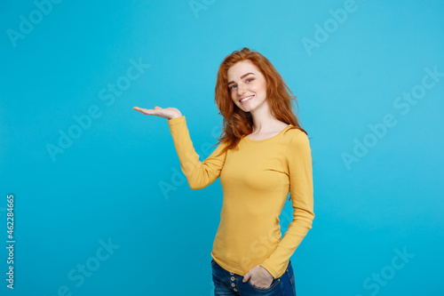 Fun and People Concept - Headshot Portrait of happy ginger red hair girl with presenting hand away and happy expression. Pastel blue background. Copy Space. photo