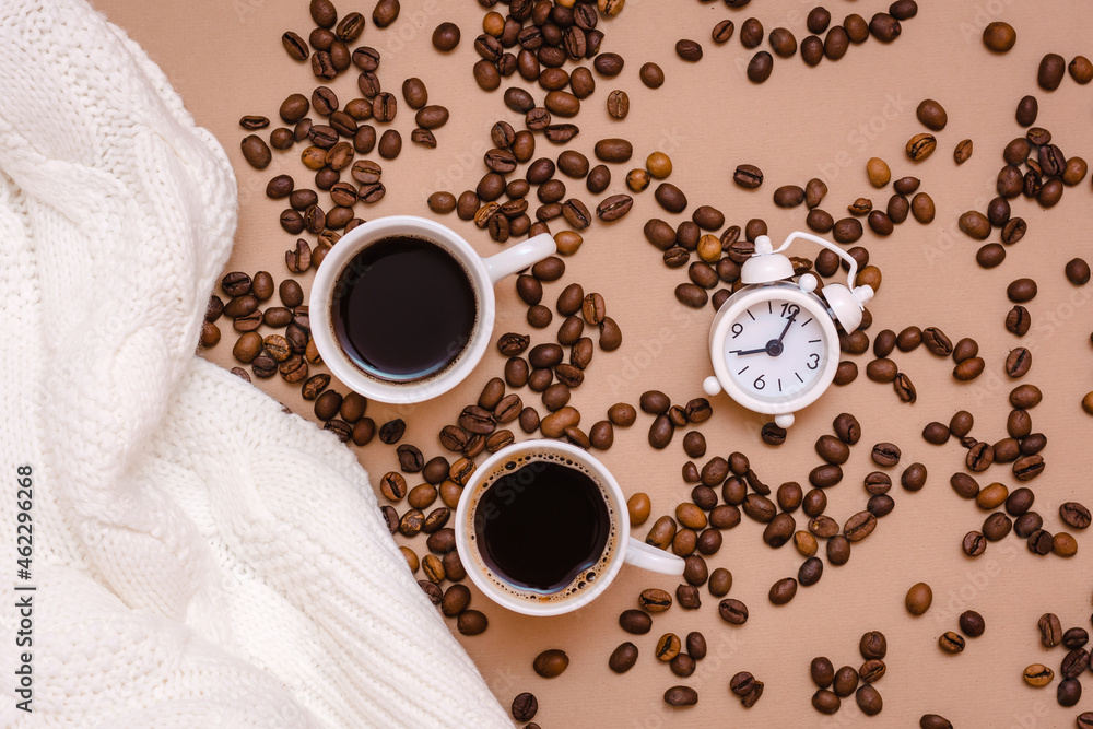 Obraz premium Two cups of black coffee, alarm clock, white sweater and coffee beans on a beige background. Cozy date and wellness. Top view