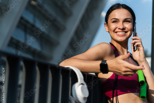 Laughing brunette woman with rejoiced face smiling wide and using mobile phone while chatting with somebody at the street after workout