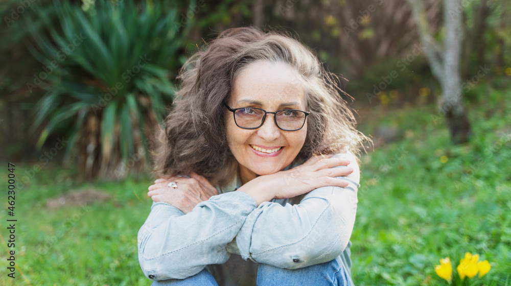 Obraz premium Portrait of a beautiful senior woman 60 years old in glasses with a charming smile looking at the camera while sitting on the grass in the garden