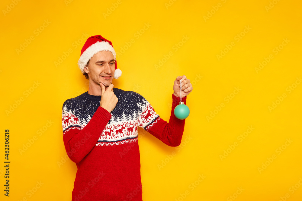 cheerful man in xmas sweater with reindeer, red Santa Claus hat holds Christmas tree toy in his hands and looks with appreciative look before buying. isolated yellow background with space for text cheerful man in xmas sweater with reindeer, red Santa Claus hat holds Christmas tree toy in his hands and looks with appreciative look before buying. isolated yellow background with space for text