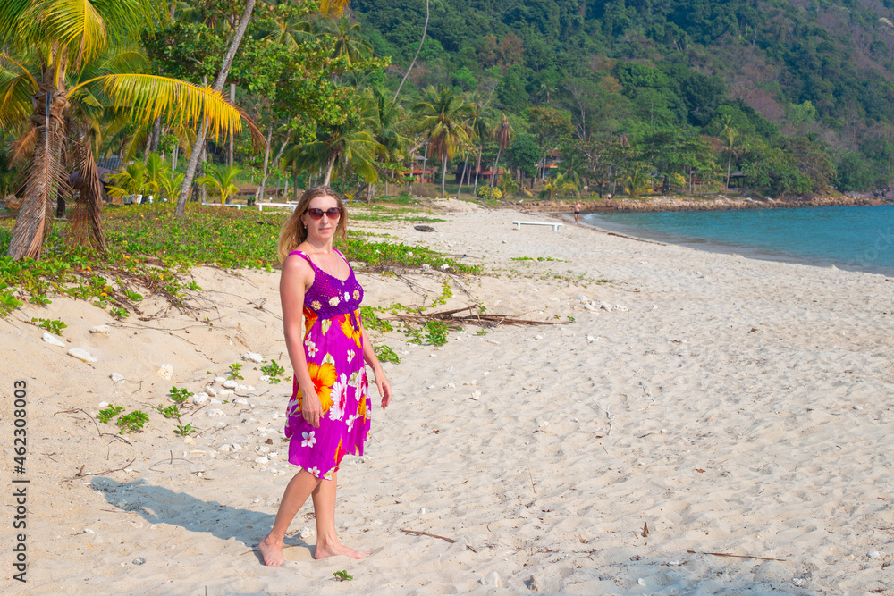 A mature woman with long blond hair walks along a sandy beach among palm trees on a tropical island in Thailand. A mature woman with long blond hair walks along a sandy beach among palm trees on a tropical island in Thailand.